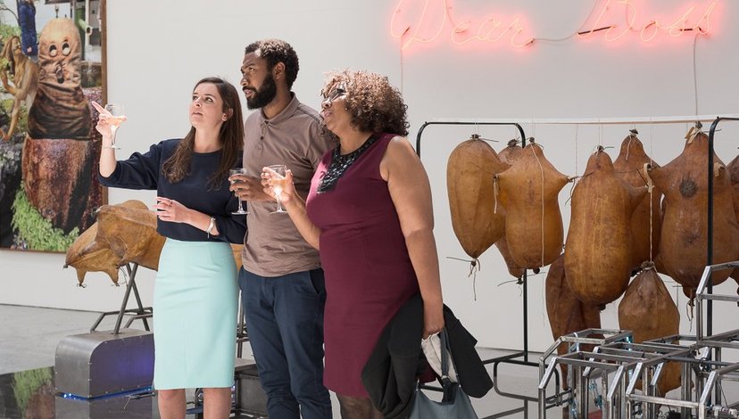 Three people discuss works in an exhibition. Neon text and inflated leather sculptures hand on the rear wall.