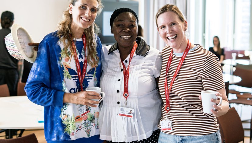 Three smiling women stand together, two holding mugs, all wearing lanyards with "Royal College of Art" visible on them.