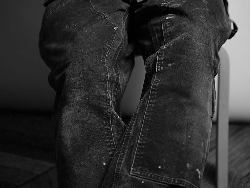 Black and white photo showing a person’s legs in worn, paint-splattered jeans, seated with knees angled inward.