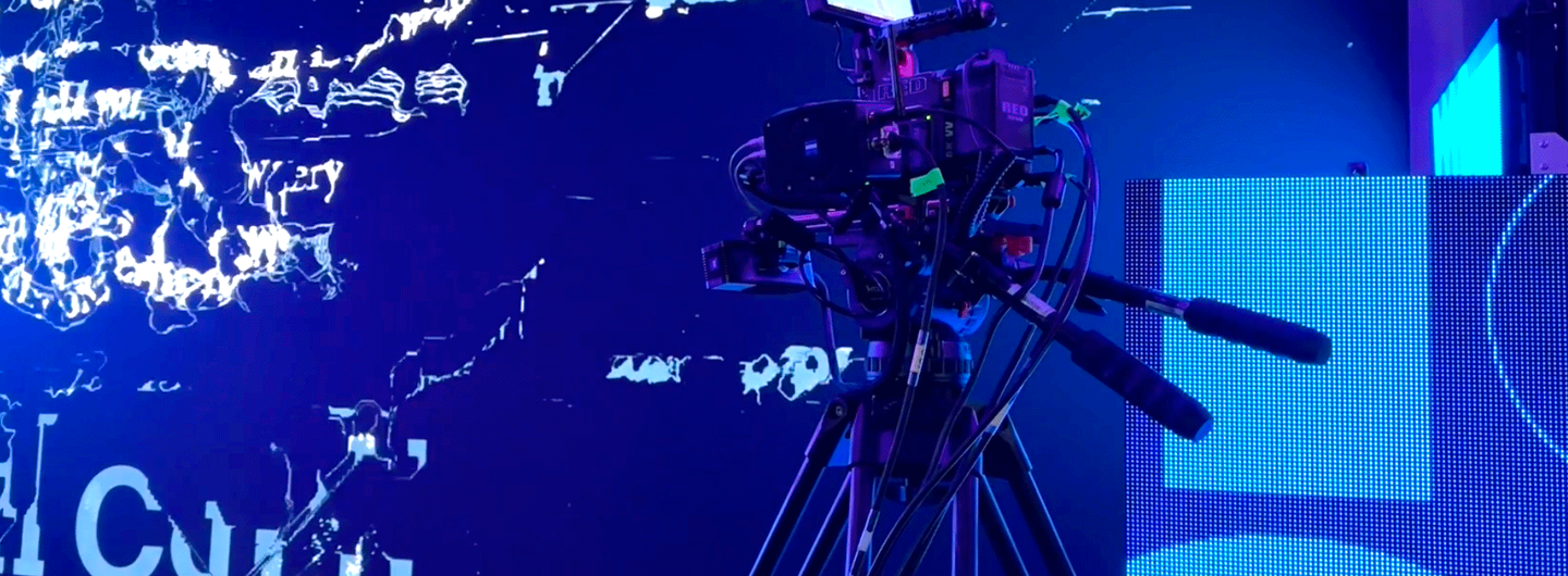 Professional video camera on tripod filming in front of a vibrant digital screen with abstract text and graphics.