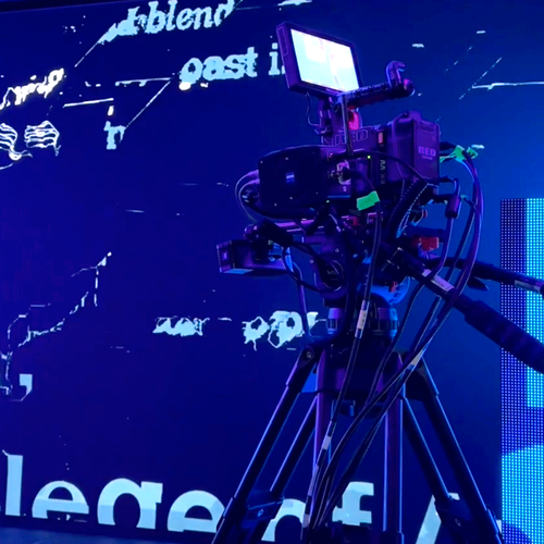 Professional video camera on tripod filming in front of a vibrant digital screen with abstract text and graphics.
